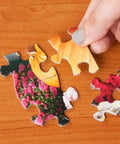 Hand holding pieces of 300 Piece Hot Air Balloon Festival Puzzle For Adults By Michael David Ward on wooden table, colorful artwork.