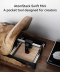 AtomStack Swift Mini laser engraver mounted on aluminum rails; side view beside a brown backpack on a desk