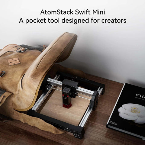 AtomStack Swift Mini laser engraver mounted on aluminum rails; side view beside a brown backpack on a desk