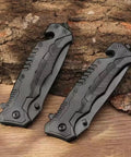 Two Black Serrated Folding Knife With Dual Edge Design For Outdoor Camping, Fishing And Hunting resting on a wooden surface.