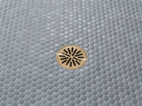 Smooth Draining Four Inch Round Shower Floor Drain With Removable Grate And Solid Gasket For Zero Fuss Tile Installation Featuring Fast Height Adjustment Plus Warm Brushed Gold Finish installed in light grey pebble tile.