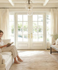 Bright living room with white French doors, beige curtains, and a woman reading on a cream sofa.