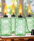 Outdoor Table Top Torch Set With 12 Oz Refill Canisters And Wicks Green Patio Torches For Luau Parties And Garden Decor DIY Backyard Lighting Includes Covers 4 Pack For A Lit Backyard Nighttime Vibe, showing green glass torches on a wooden table, lit flames.