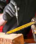 Silver precision oiler pen applicator with knurled cap held by gloved hand, near a yellow chainsaw bar.