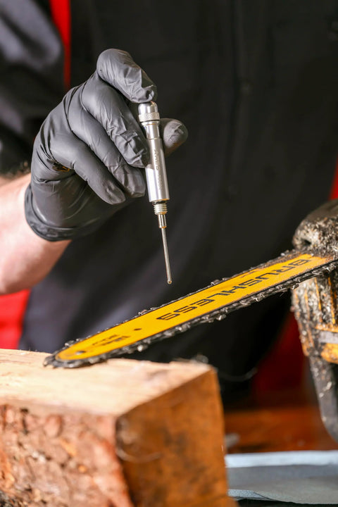 Silver precision oiler pen applicator with knurled cap held by gloved hand, near a yellow chainsaw bar.