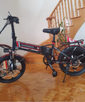 Black folding electric bike with red accents on wooden floor near staircase.