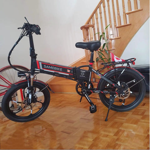 Black folding electric bike with red accents on wooden floor near staircase.
