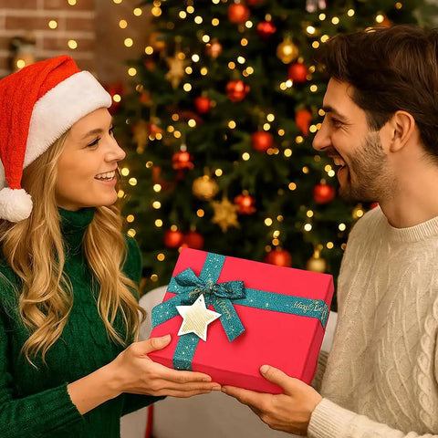 Two people exchanging a red gift box with teal ribbon in front of a decorated Christmas tree.