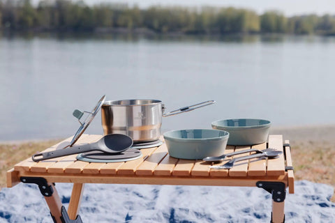 Wildfare Go 14 Piece Stainless Steel Camp Kitchen Set with 1.65 Quart Saucepan, bowls, utensils on a wooden table by the lake.