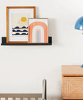 Damage Free Picture Ledge 21 Inch Floating Shelf With Adhesive Strips No Tools Needed Holds Photos And Decorations Up To 5 Pounds Slate Finish includes 1 ledge and 10 strips holding framed art above a wooden cabinet with a blue lamp.