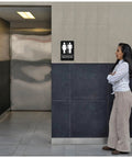 Woman standing near ADA Braille Unisex Bathroom Sign With Double Sided 3M Tape ABS Plastic Gender Neutral Restroom Sign For Offices And Restaurants 9 By 6 Inches Sleek Modern Mountable Office Sign For Biz Spaces on wall beside restroom door.