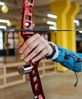 Archer holding a red metallic bow in an indoor range, focusing on the hand grip and arrow alignment.