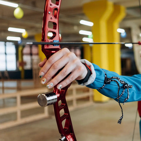 Archer holding a red metallic bow in an indoor range, focusing on the hand grip and arrow alignment.