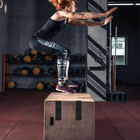 Woman performing a jump exercise on a wooden box in gym with fitness equipment in the background.