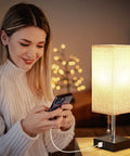 Woman using phone by lit Set Of 2 USB Nightstand Lamps With Grey Fabric Shade Small Table Lamps For Bedroom End Table Living Room Or Home Office Built In 5 Volt 2 Amp USB Ports To Charge Two Devices While You Sleep