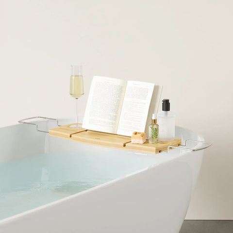 Aquala Extendable Bathtub Tray Caddy With Book Stand And Wine Glass Holder Phone Stand Foldaway Bar For Books Or Tablets Razor Loofah Scrubber Hooks Natural Finish over a bathtub with book and wine glass visible.