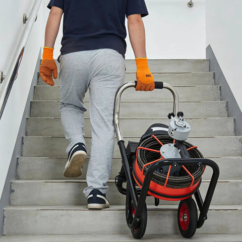 Person carrying a 450W Auto Feed Drain Auger with 100 Ft steel cable up stairs, ready for tackling nasty clogs.