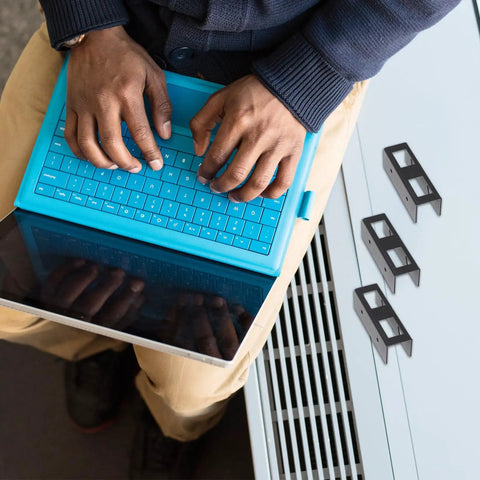 Person using a blue tablet on their lap with hands on keyboard next to Heavy Duty Laptop Stand And Tablet Mount Under Desk Organizer Rack Black.