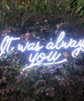 Neon sign on foliage stating 'It was always you' in white script lighting up a garden setting.