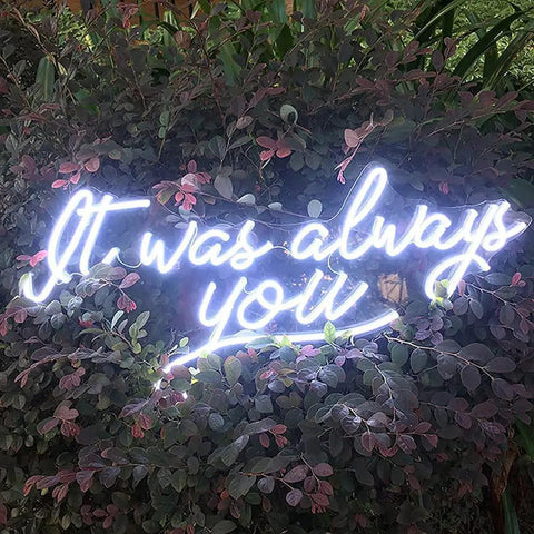 Neon sign on foliage stating 'It was always you' in white script lighting up a garden setting.