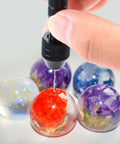 Pin vise hand drill with black grip drilling a red resin globe; assorted colored resin spheres in background