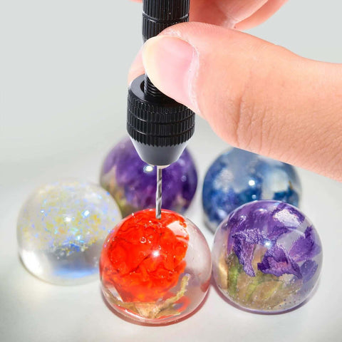 Pin vise hand drill with black grip drilling a red resin globe; assorted colored resin spheres in background