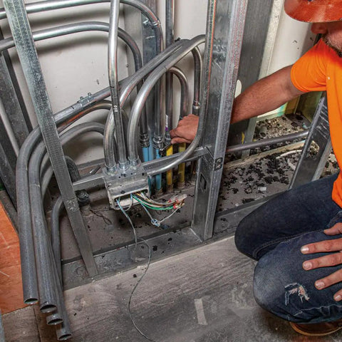 Person working on metal pipe connections in a building wall, surrounded by electrical cables and metal studs.