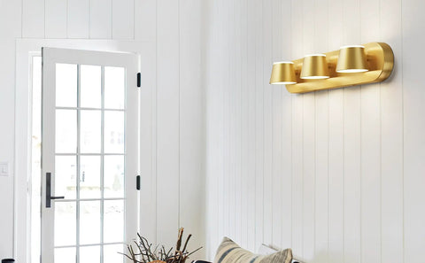 Gold three-light bathroom fixture on white wall above a door, showcasing its modern design with cylindrical shades.