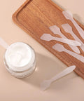 300 Pack Mini Cosmetic Spatulas 3.2 By 0.6 Inches Disposable Plastic Spatulas Clear For Facial Creams And Skin Care, No Cross Contamination, Great For Serums, Moisturizers, Face Masks, And Spa Day Vibes. Clear plastic cosmetic spatulas beside cream jar and wooden tray