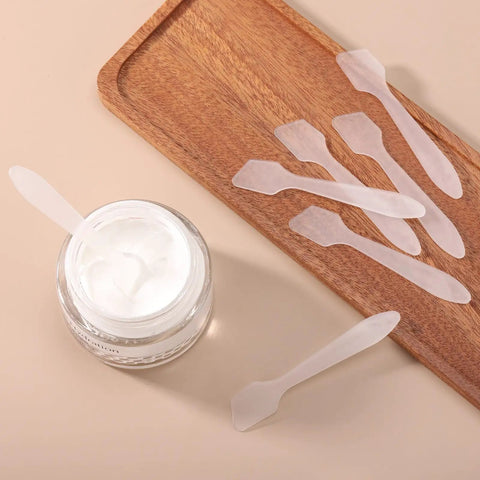 300 Pack Mini Cosmetic Spatulas 3.2 By 0.6 Inches Disposable Plastic Spatulas Clear For Facial Creams And Skin Care, No Cross Contamination, Great For Serums, Moisturizers, Face Masks, And Spa Day Vibes. Clear plastic cosmetic spatulas beside cream jar and wooden tray