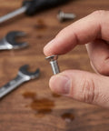 Close-up of a hand holding a M8x60mm stainless steel flat head hex socket screw, showing full thread detail, tools in background.