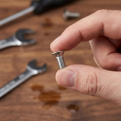 Close-up of a hand holding a M8x60mm stainless steel flat head hex socket screw, showing full thread detail, tools in background.