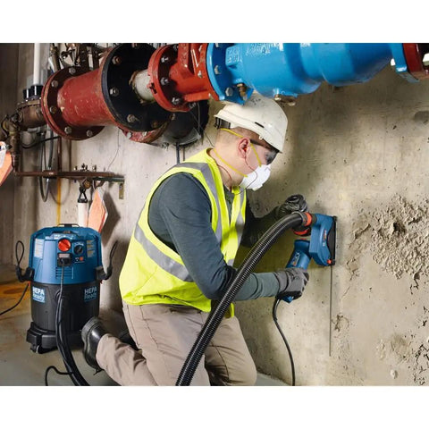 Worker using power tool with vacuum under pipes, wearing safety gear and mask, onsite construction environment