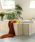 Bathroom with a white tub, beige tiled surround, an orange towel, and decorative plants.