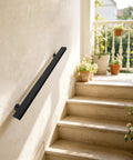 Badass Black Stainless Steel Wall Mount Handrail For Stairs And Hallways With Heavy Duty Support and Tight Secure Grip For Indoor Renovation And Exterior Installations mounted on a light wall by stairs.