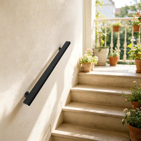 Badass Black Stainless Steel Wall Mount Handrail For Stairs And Hallways With Heavy Duty Support and Tight Secure Grip For Indoor Renovation And Exterior Installations mounted on a light wall by stairs.