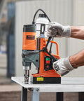 Front view of orange VEVOR magnetic drill press mounted on a metal table, with a gloved hand operating the handle.