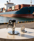 Heavy Duty Marine Grade 316 Stainless Steel Door Holder Catch With Rubber Bumper To Stop Slamming Noise And Keep Your RV Or Yacht Door Securely Locked In Place Without Rusting In Saltwater Conditions on a table with a cargo ship in the background.