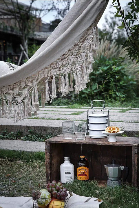 Double Outdoor Cotton Hammock With Fringes For Two Adults Perfect For Beach Or Patio Chill Sessions Camping Or Travel 58 Inch Wide Supports Up To 400 Lbs Brazilian Style Portable Canvas Bed For Relaxing with visible fringes in a garden setting.