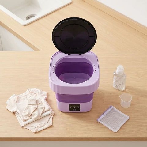 Slick Mini Folding Washing Machine 6 Liter Portable Unit on wooden table with baby clothes, detergent, and measuring cup.