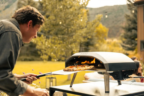 Koda 16 Inch Propane Gas Pizza Oven with 28 MBAR Regulator and Hose, outdoor setup with pizza being cooked, Foundry Black.