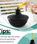 Close-up of a lit black metal table top torch on a white table, part of a Six Pack Metal Table Top Torches With 23.5 Oz Citronella Fuel For Outdoor Parties And Garden Decor Refillable Flame Torches With Wicks And Covers Long Lasting Burn Time Of Over 18 Hours set.