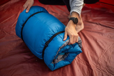 Person securing blue sleeping bag with 'Two Pack 48 Inch Sleeping Bag Straps With Quick Release Buckles For Universal Fit' in red tent.