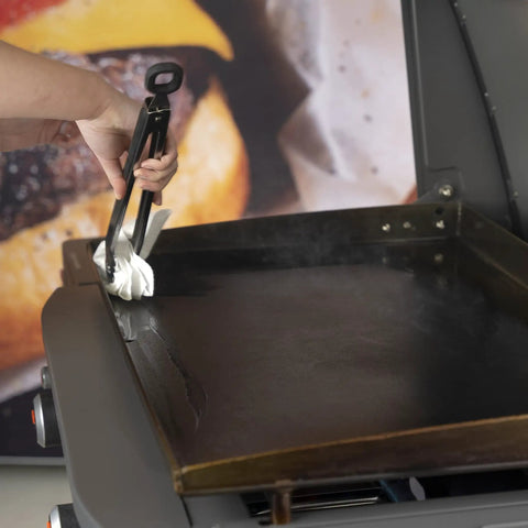 Hand using tongs with cloth to apply Griddle Seasoning And Cast Iron Conditioner 2 In 1 Formula For Nonstick Release And Rust Protection
