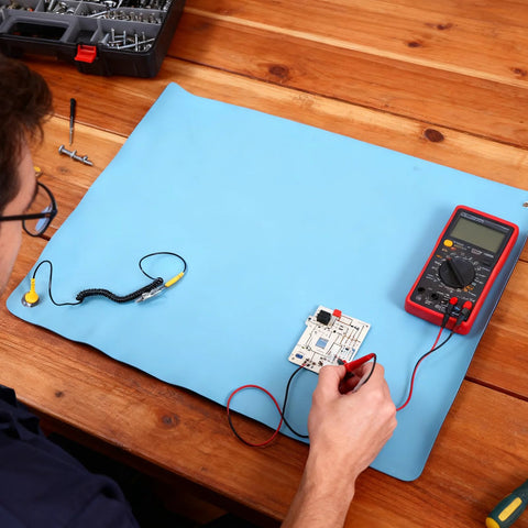 Blue anti-static ESD mat on wooden desk with PCB board, grounding wire, and red digital multimeter; top-down view.