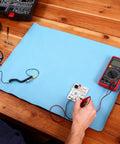 Blue anti-static ESD rubber mat on a wooden workbench with grounding wire, PCB, and red digital multimeter.