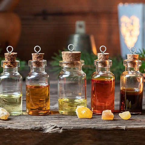 Five glass bottles with corks filled with various colored oils on a wooden surface, with amber stones in front and a heart-shaped light in background.
