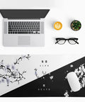 Top view of an extended black and white Sakura cherry blossom desk pad for gaming office or home setup, 31.5 by 11.8 inches, non-slip rubber base, stitched edges, with a laptop, mouse, coffee, glasses, and plant displayed.