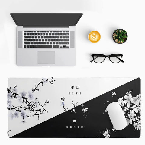 Top view of an extended black and white Sakura cherry blossom desk pad for gaming office or home setup, 31.5 by 11.8 inches, non-slip rubber base, stitched edges, with a laptop, mouse, coffee, glasses, and plant displayed.