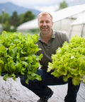 Gardener holding leafy greens outdoors in a greenhouse environment, showcasing fresh produce.
